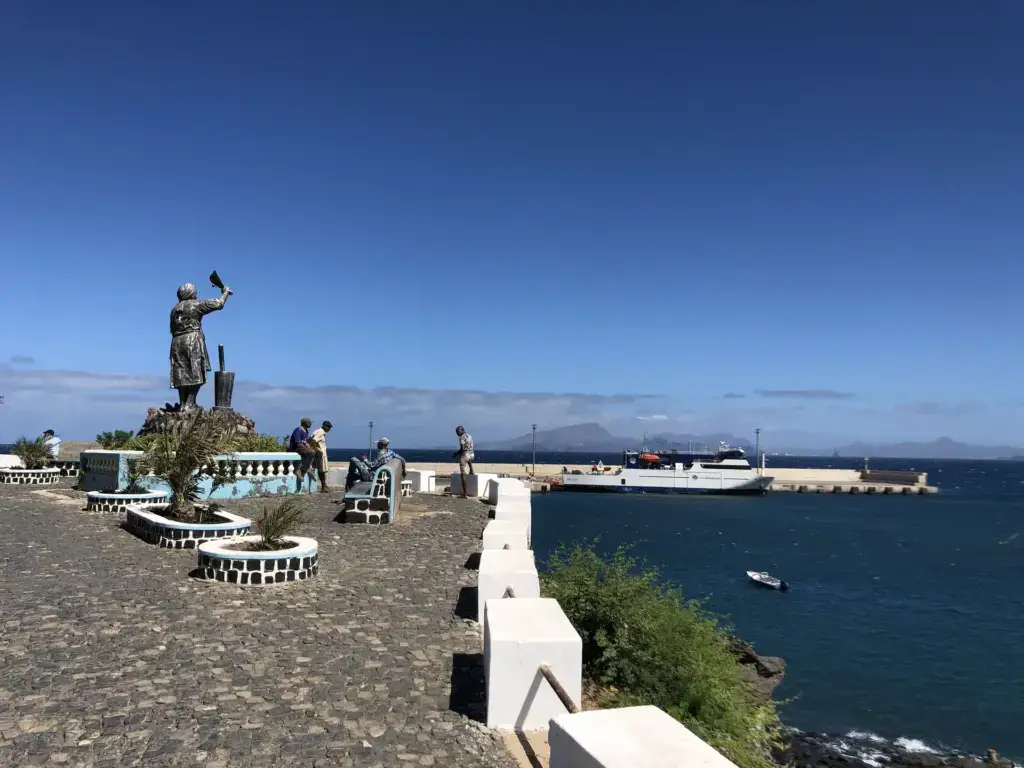 Statue of a Cape Verdean woman on the cobbled waterfront at Porto Novo, Santo Antão, with the ferry from São Vicente docked behind.