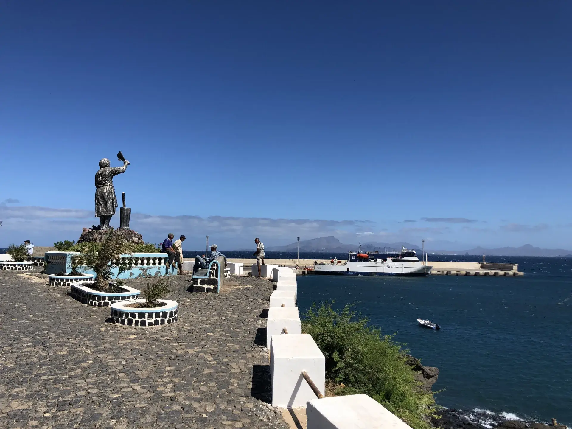 Statue of a Cape Verdean woman on the cobbled waterfront at Porto Novo, Santo Antão, with the ferry from São Vicente docked behind.